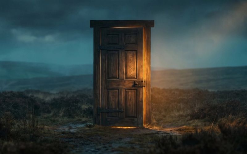 A heavy panelled door standing shut and completely alone on open moorland at dusk, warm light blazing around its edges, with open ground running past both sides of the frame