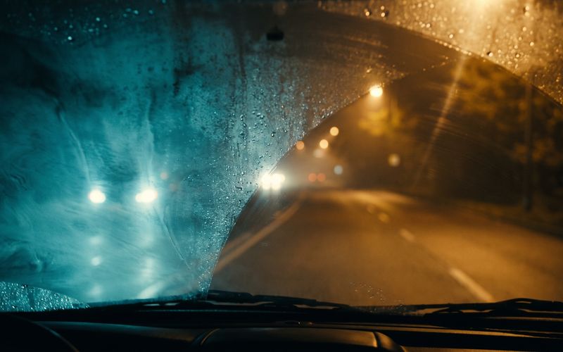A windshield fogged across most of its surface with one clear patch, warm amber lights showing through only where the glass is clear