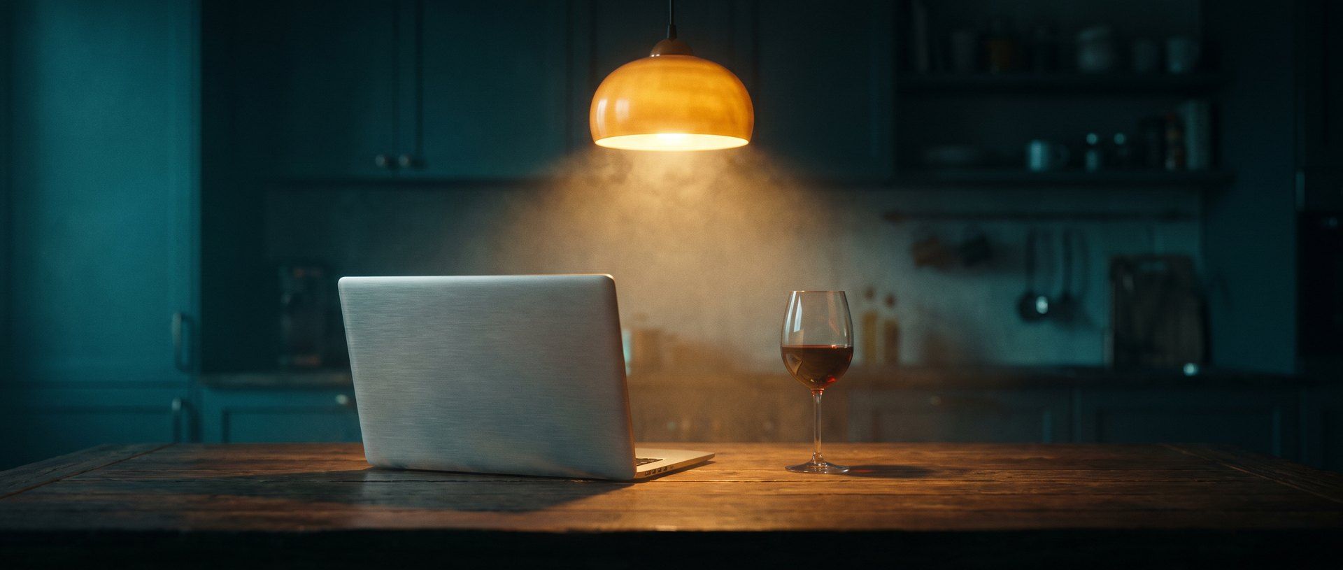 A closed laptop and a poured glass on a kitchen table at night under a warm pendant lamp