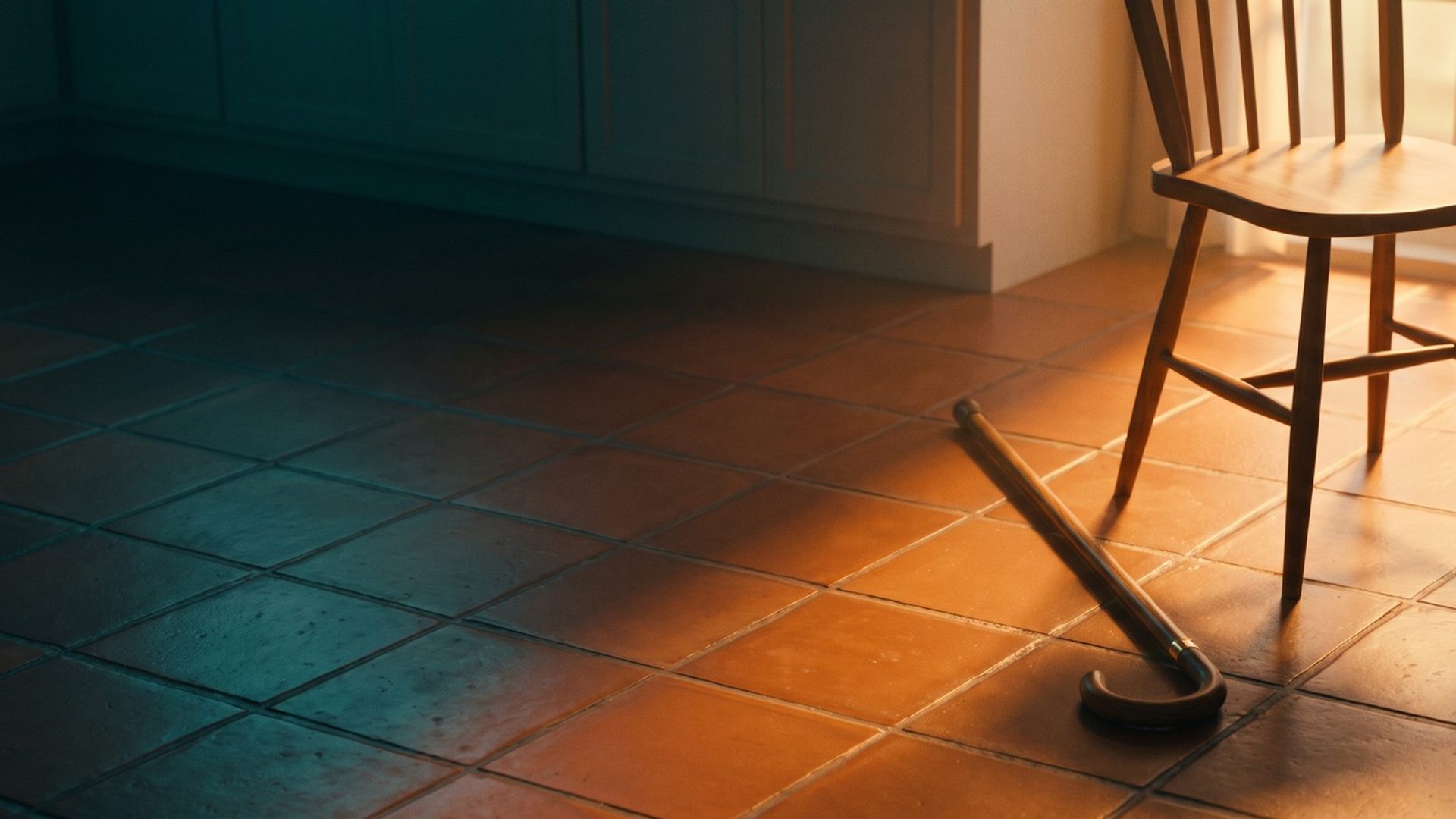 A wooden walking cane lying on a terracotta kitchen floor in low morning sun, beside a chair pushed back from a small table with a coffee cup still on it