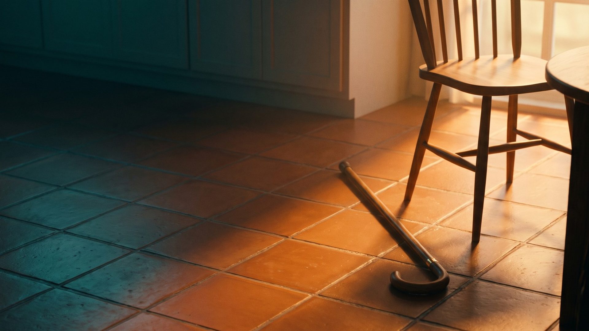 A wooden walking cane lying on a terracotta kitchen floor in low morning sun, beside a chair pushed back from a small table with a coffee cup still on it