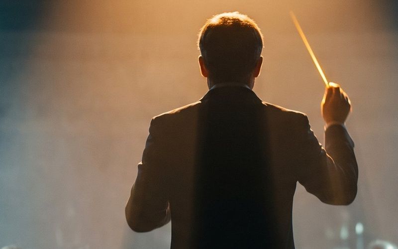 A conductor seen from behind, baton raised under a warm spotlight, leading an orchestra through stage haze.