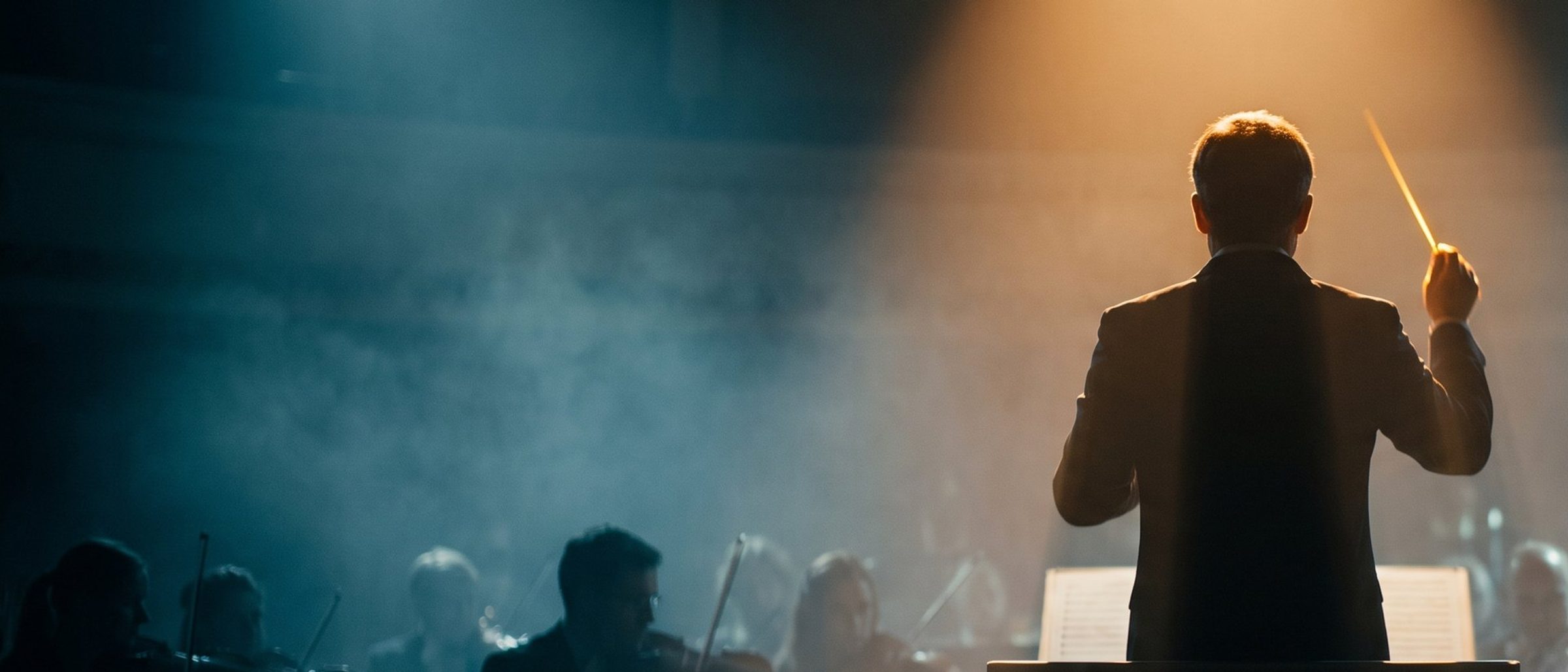 A conductor seen from behind, baton raised under a warm spotlight, leading an orchestra through stage haze.