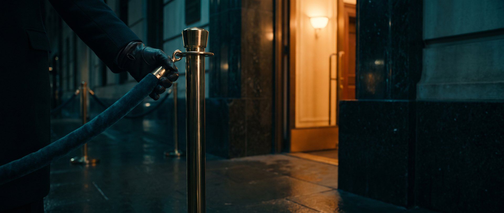 A gloved hand unhooking a velvet rope from a stanchion and holding it aside at a lit entrance