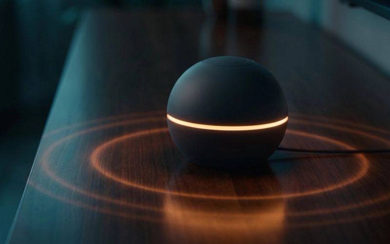 A black smart speaker with a glowing amber ring sitting on a dark wooden table, rings of light spreading across the surface around it.