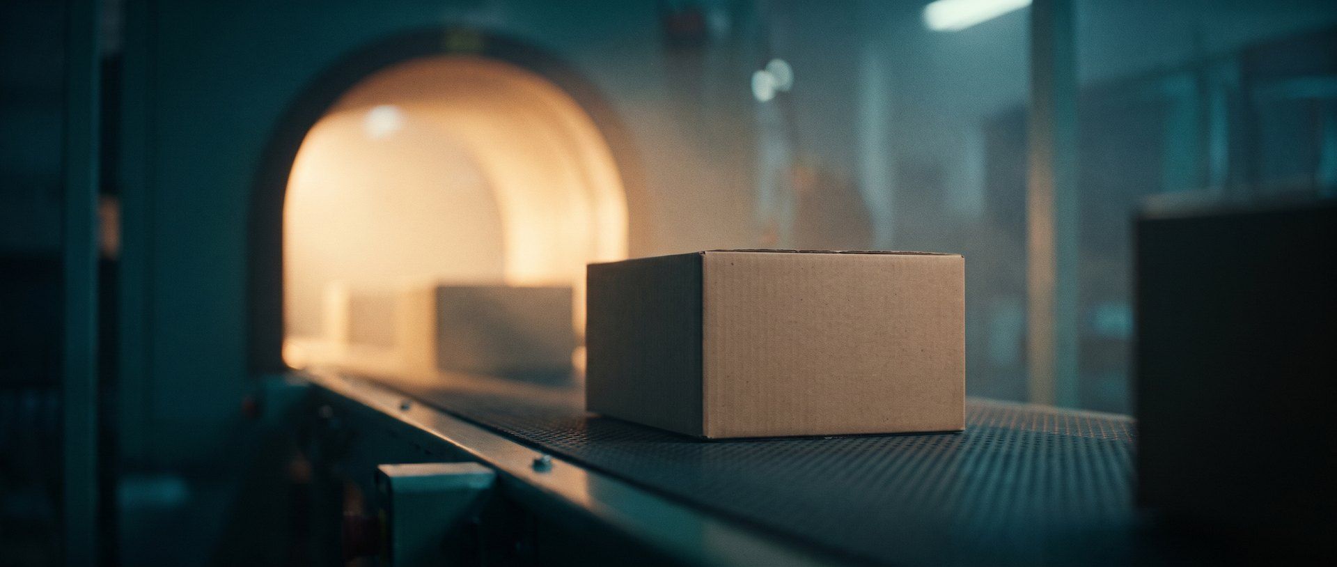 A plain cardboard shipping box riding a dark conveyor belt toward a glowing amber archway in a teal-lit fulfillment center, more boxes following behind it.