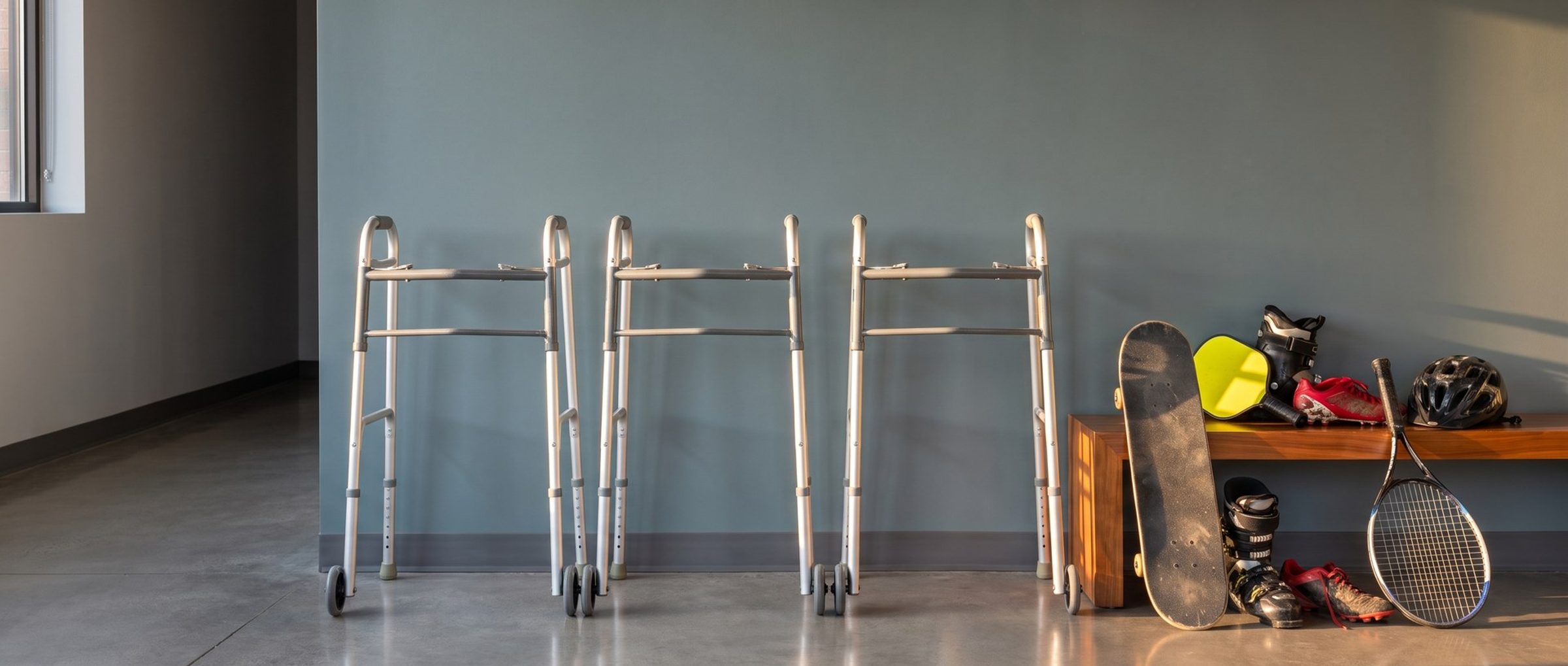 Three identical aluminum walkers standing in a neat row against a teal-gray clinic wall, beside a wooden bench piled with a skateboard, a pickleball paddle, ski boots, soccer cleats, a bike helmet and a tennis racket