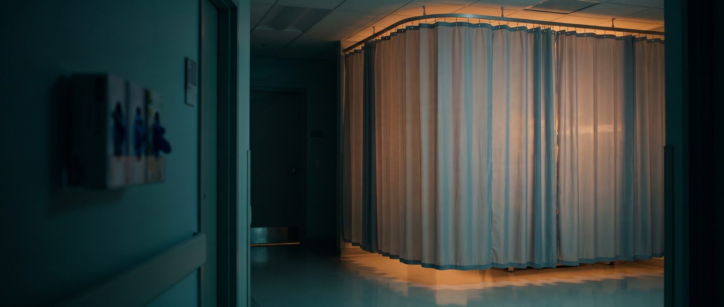 A pale privacy curtain drawn closed on its ceiling track around an exam bay, warm light glowing through the fabric and spilling under the hem, the clinic corridor dim beside it