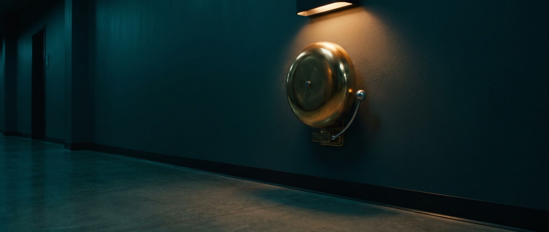 A brass alarm bell mounted high on the wall of an empty corridor, lit by a single warm lamp