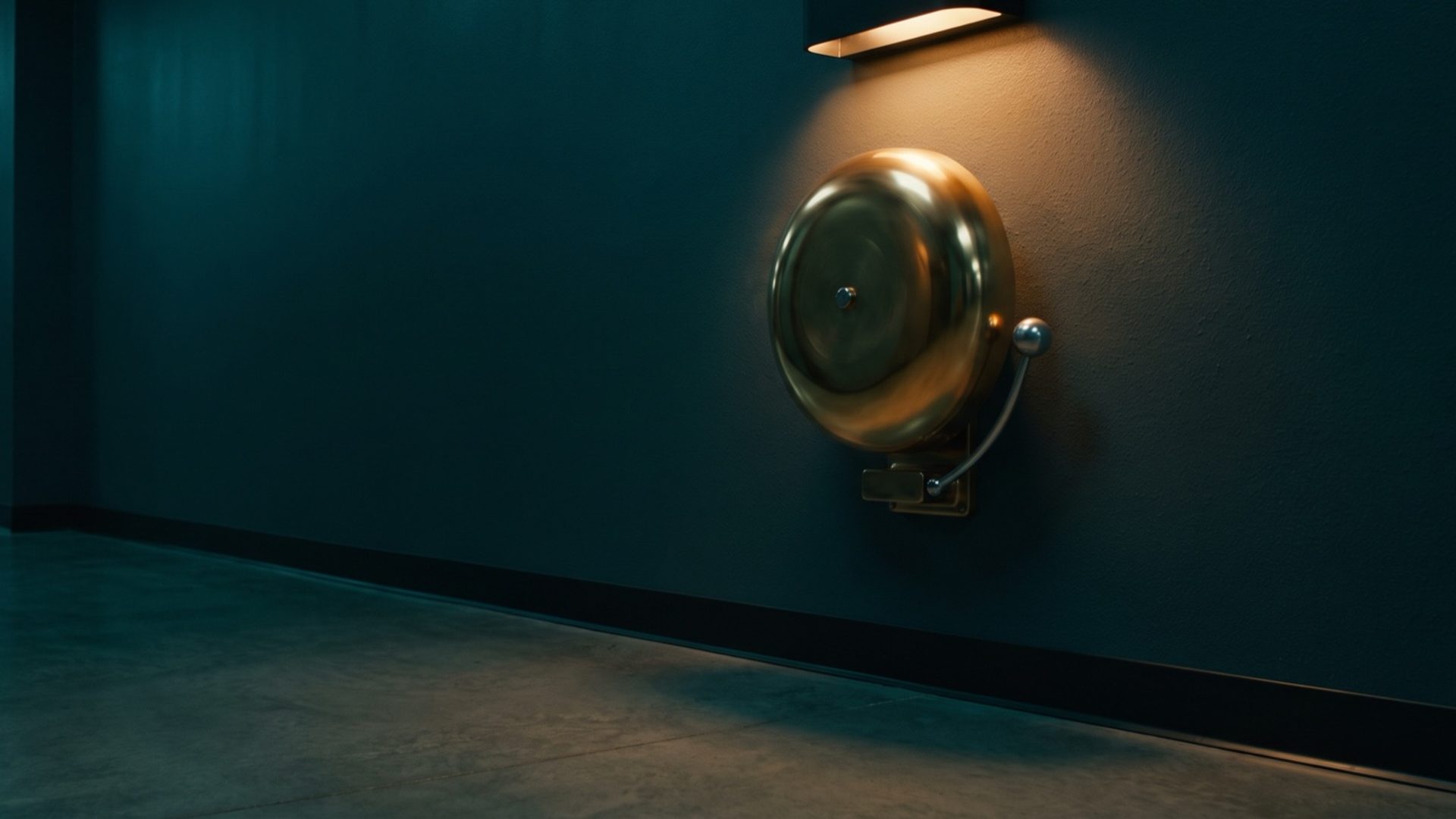 A brass alarm bell mounted high on the wall of an empty corridor, lit by a single warm lamp