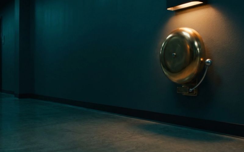 A brass alarm bell mounted high on the wall of an empty corridor, lit by a single warm lamp