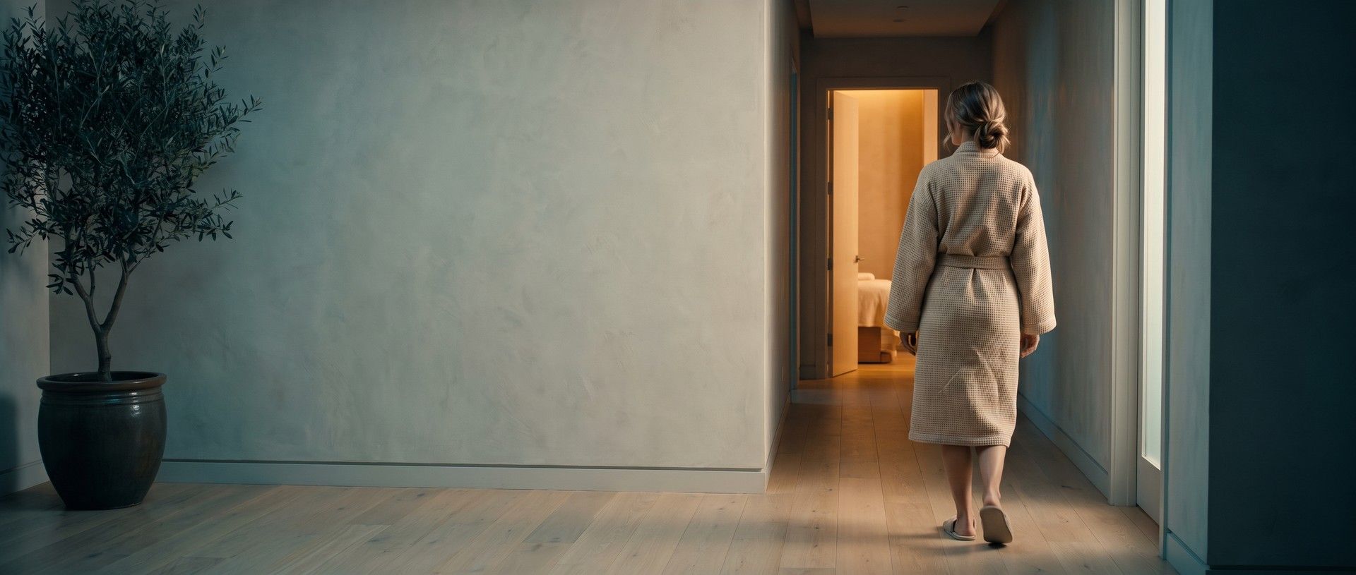 A client seen from behind in a waffle-knit robe and slippers walking down a calm med spa hallway toward an open treatment room glowing with warm light