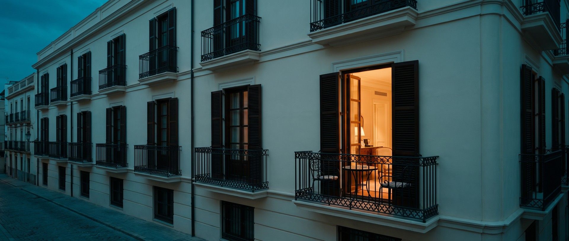 A cream historic hotel facade at blue hour with a row of dark wrought-iron balconies and shuttered windows, and one balcony lit warm, its French doors open behind two bistro chairs and a small round table