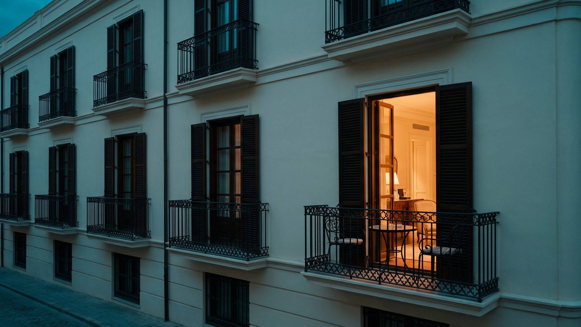A cream historic hotel facade at blue hour with a row of dark wrought-iron balconies and shuttered windows, and one balcony lit warm, its French doors open behind two bistro chairs and a small round table
