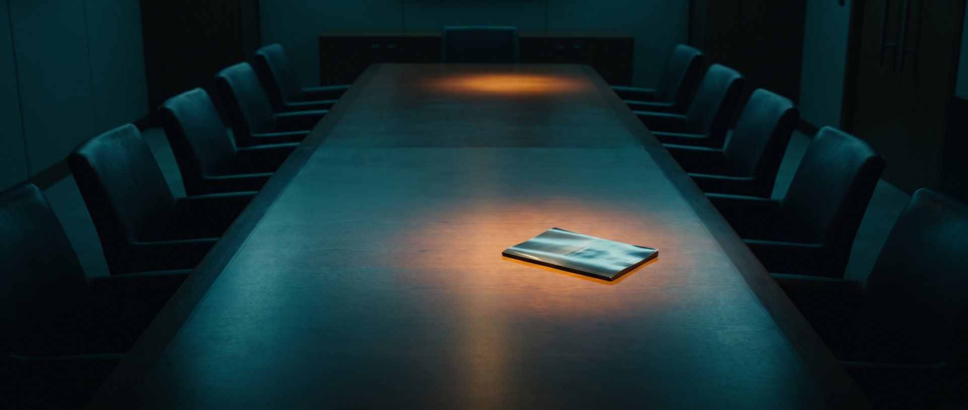 A single slim brochure lying alone under a pool of warm light on a long dark boardroom table set for twelve empty chairs