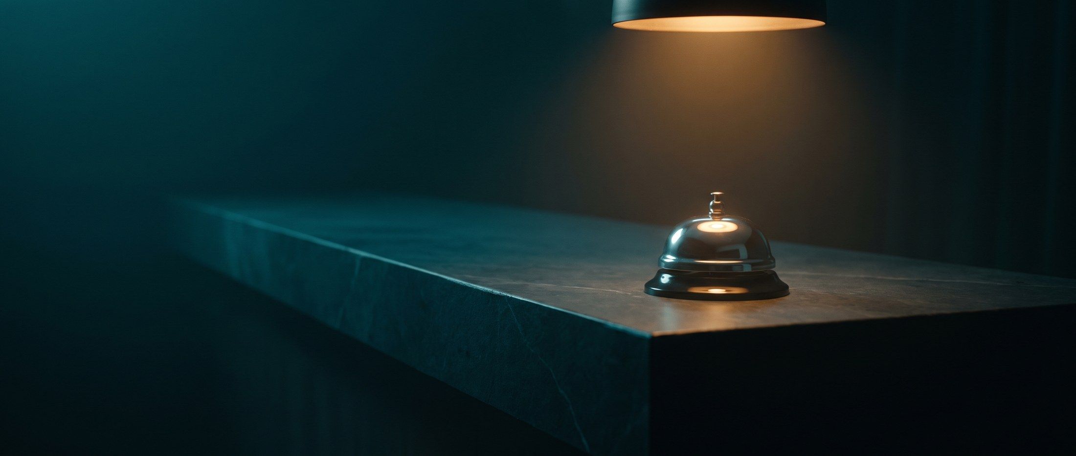 A single lit brass service bell alone on a long empty counter in a darkened room