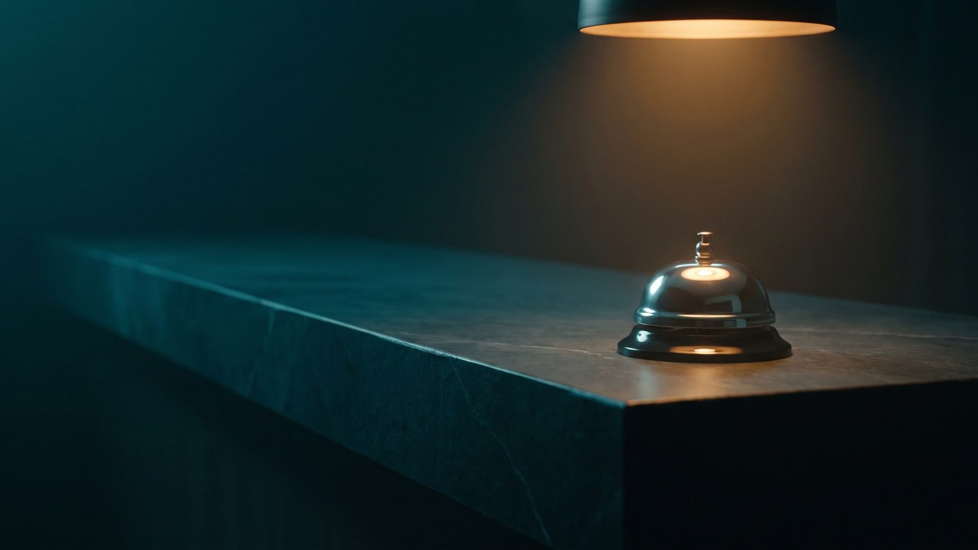 A single lit brass service bell alone on a long empty counter in a darkened room