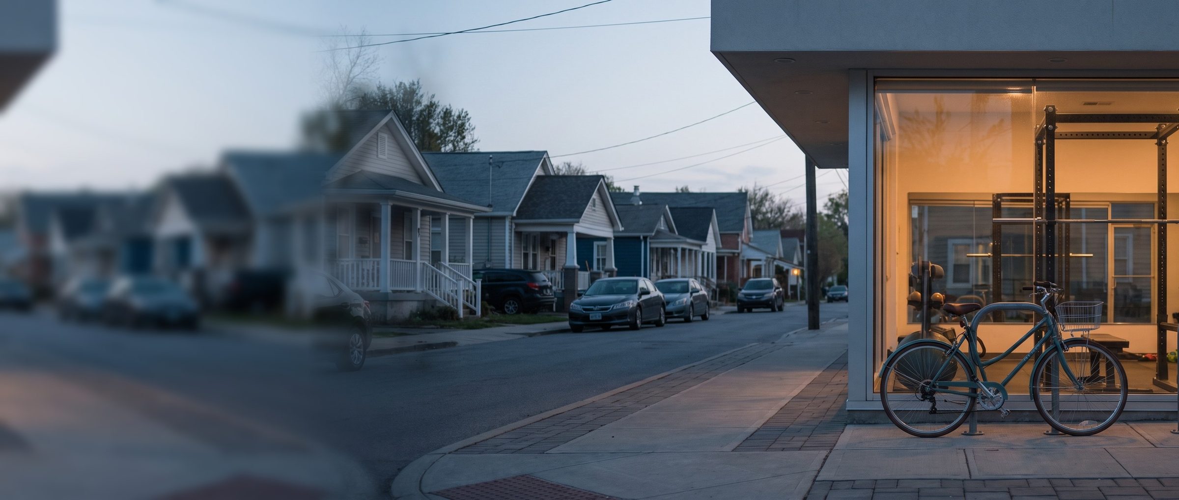 A teal commuter bicycle with a front basket on the sidewalk outside a small corner gym at dusk, its glass front glowing warm with a squat rack and dumbbells inside, a row of porch houses and parked cars down the residential street