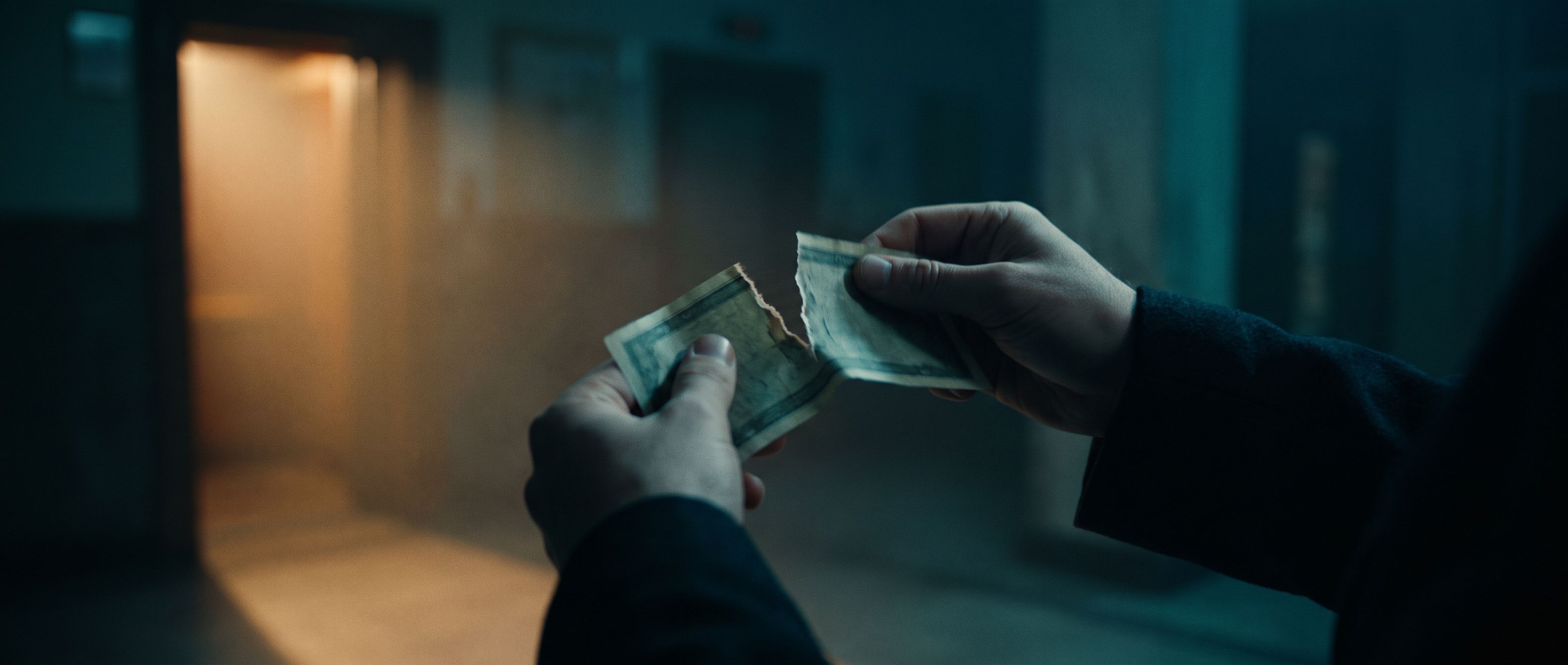 Two hands tearing a worn dollar bill in a dim hallway, warm light spilling from a doorway behind