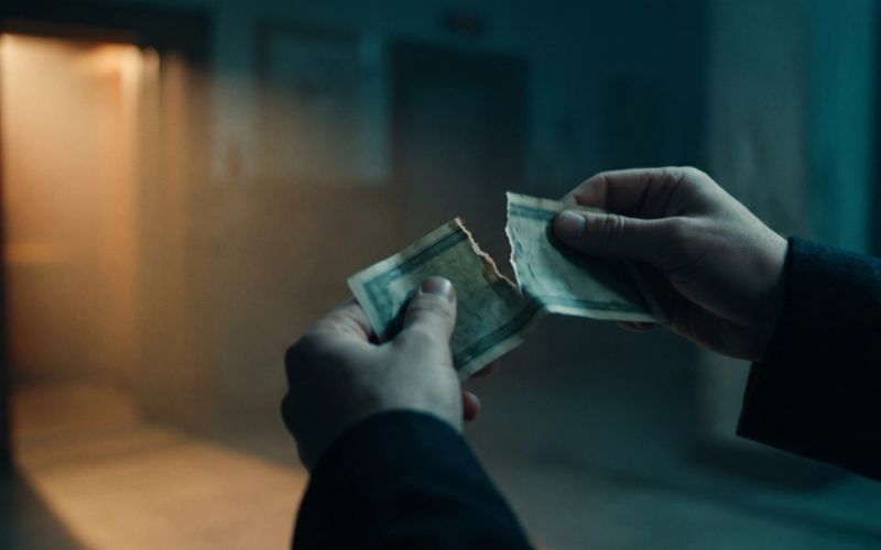 Two hands tearing a worn dollar bill in a dim hallway, warm light spilling from a doorway behind