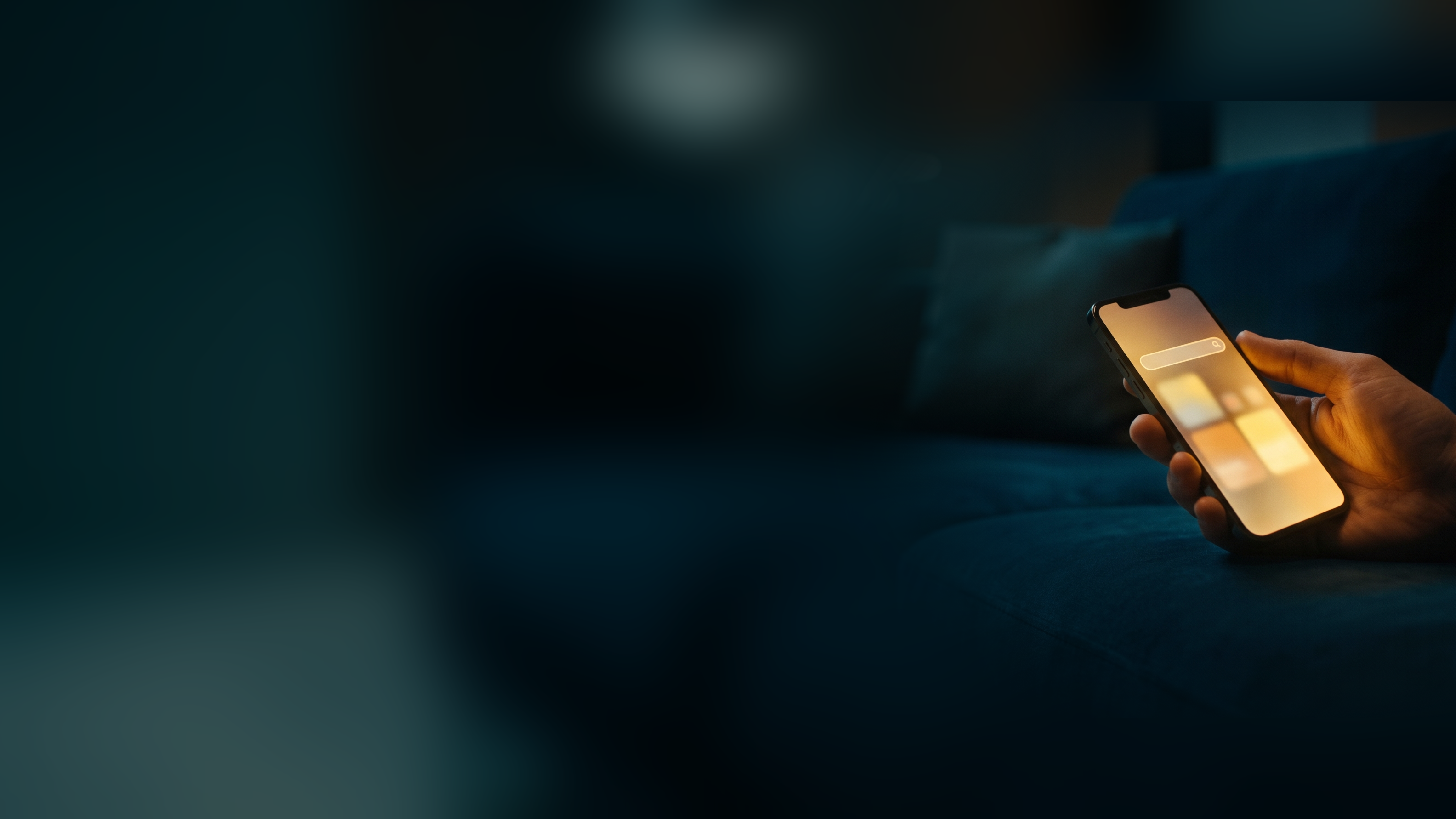 A hand holding a phone that glows with a search page on a dark living room couch at night
