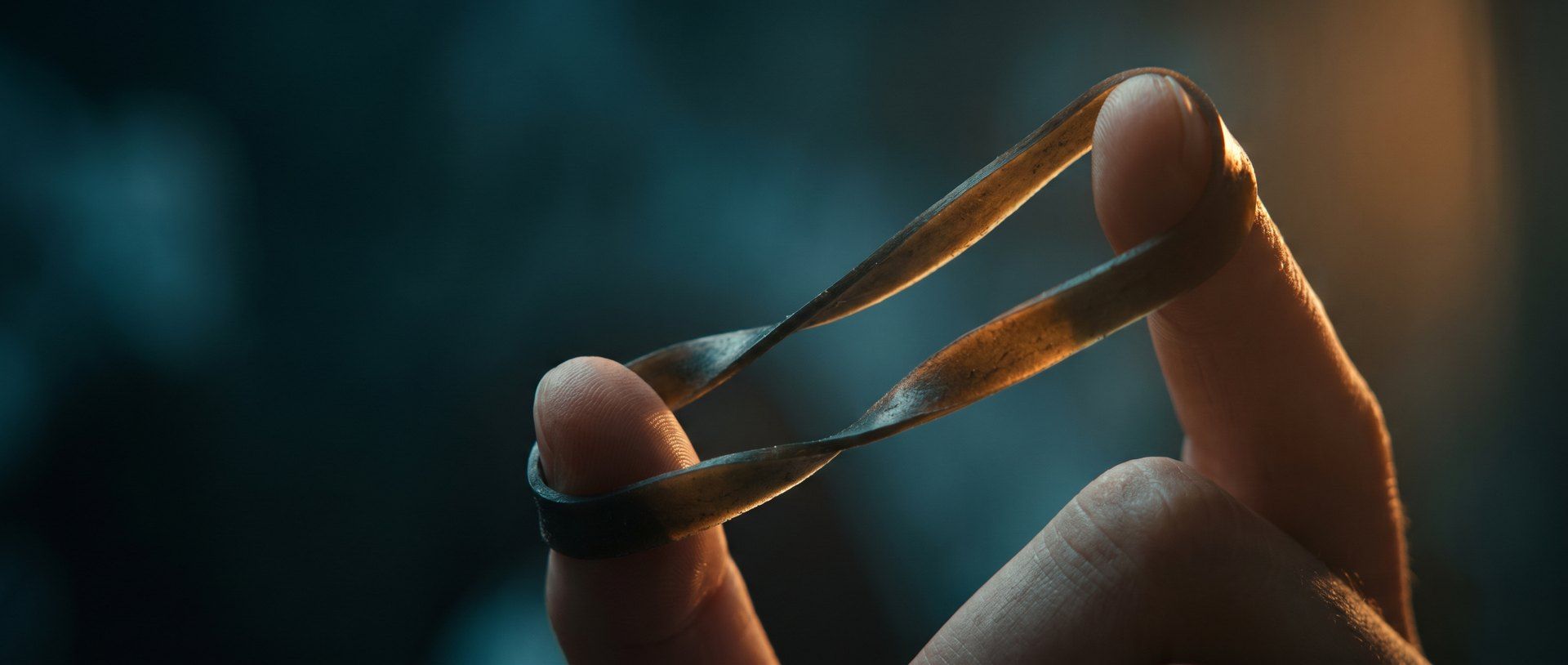 A rubber band stretched and twisted between two fingers in warm light against a dark teal background
