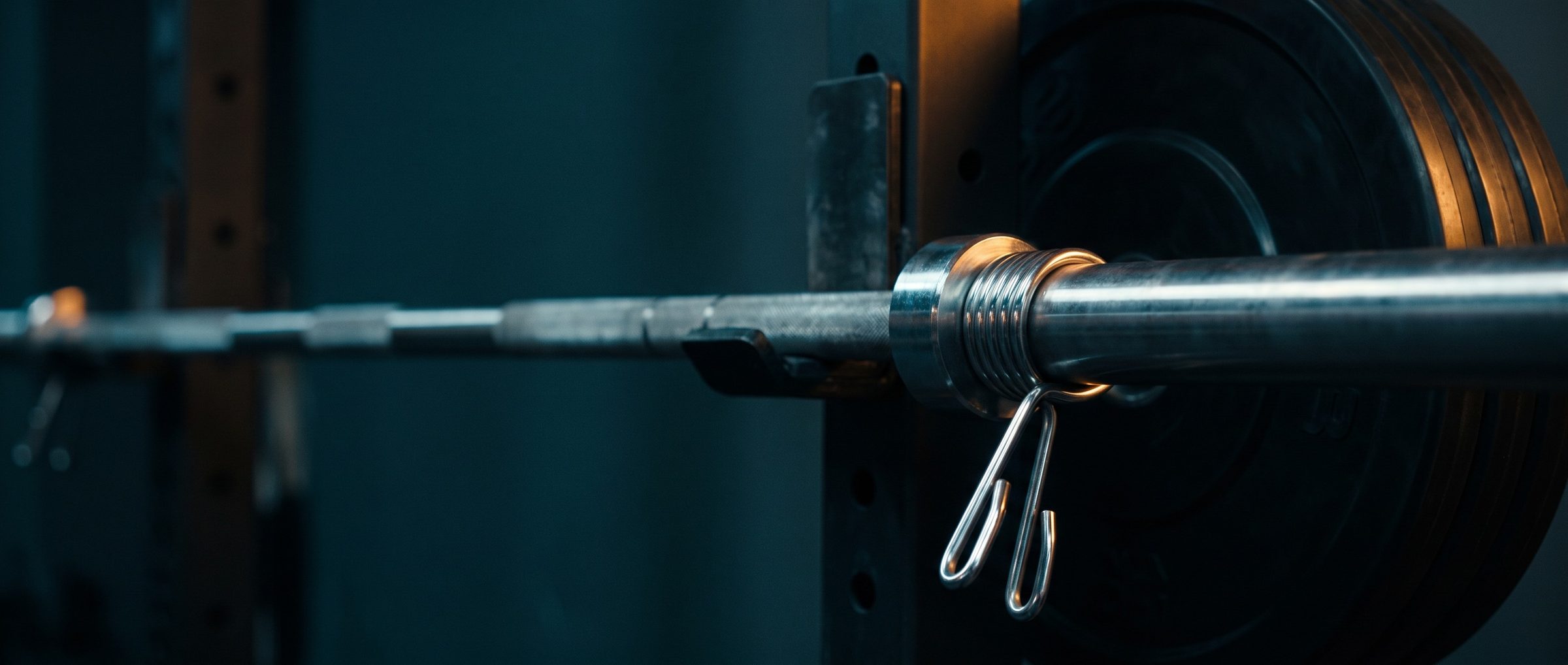 Close-up of a chrome spring collar clamped on a loaded barbell against black weight plates, its coiled spring and squeeze handles lit warm, the bar running off into a dark teal gym