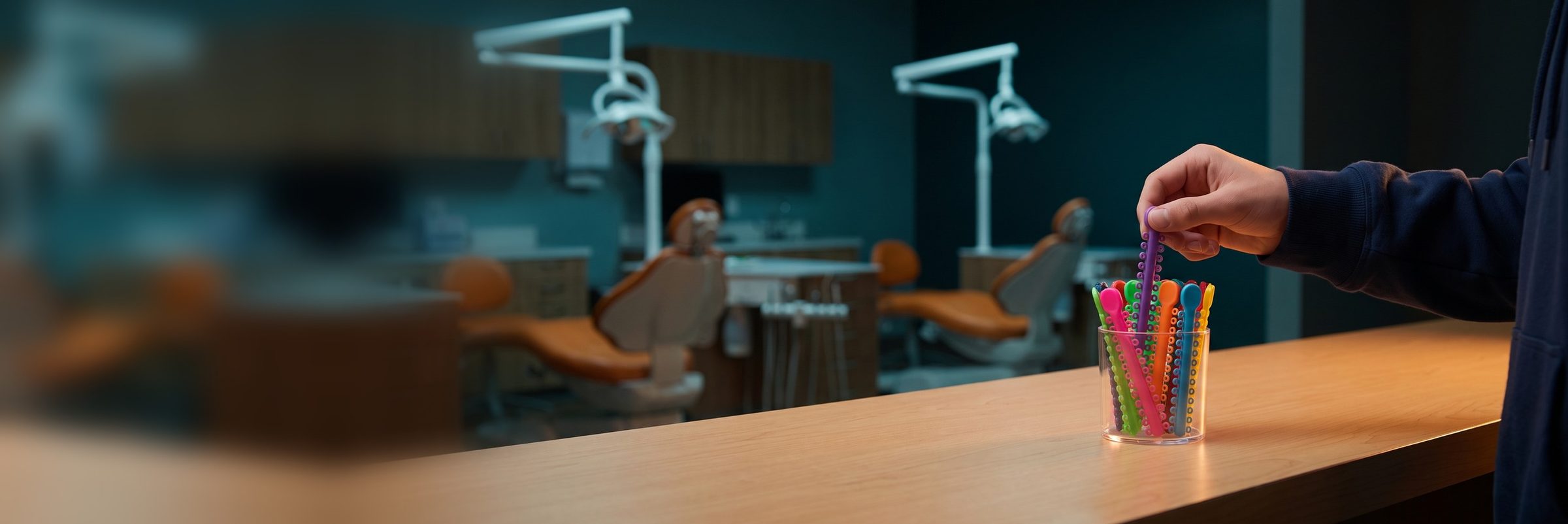 A teenager's hand pulls a purple stick of braces elastic ties from a cup of brightly colored ties on an orthodontic office front counter, with orange patient chairs blurred in the treatment area behind