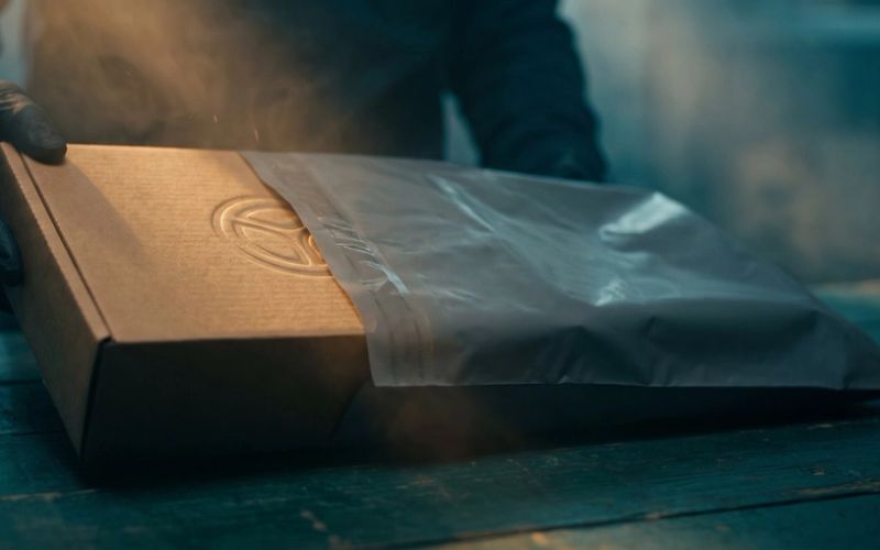 A warm kraft box with an embossed maker's emblem being slid into a plain unmarked shipping mailer, the emblem disappearing under the gray wrapper
