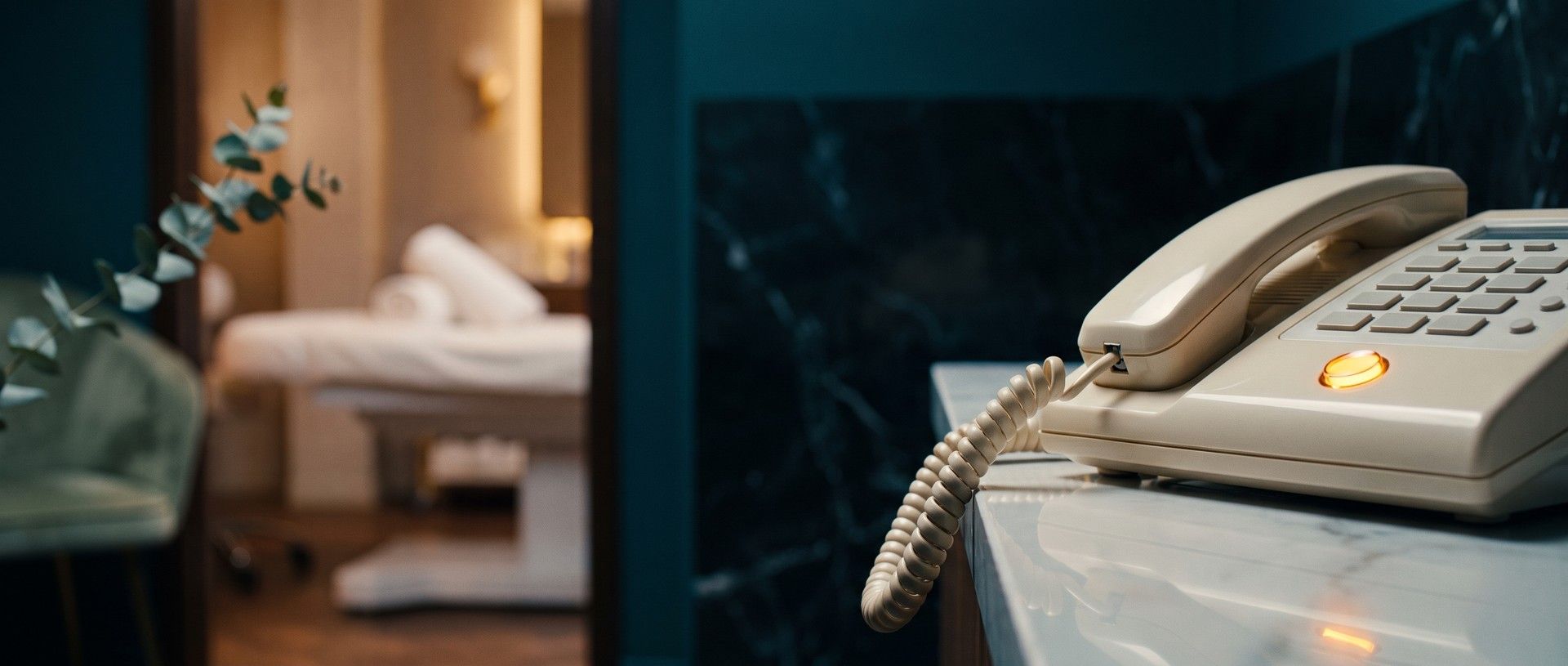 A cream desk phone on a marble counter with its line button glowing amber and the handset untouched, a med spa treatment room softly lit through the doorway behind