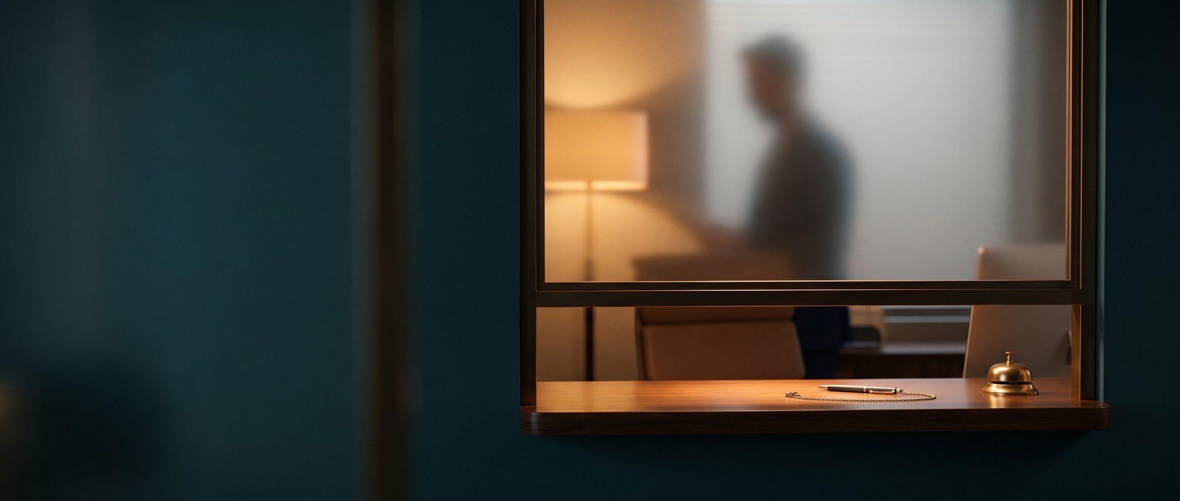A medical office check-in window seen from the waiting room: frosted privacy glass turns the staff member at a lamp-lit desk into a blurred silhouette, while a pen on a chain and a brass bell sit on the ledge below the open pane