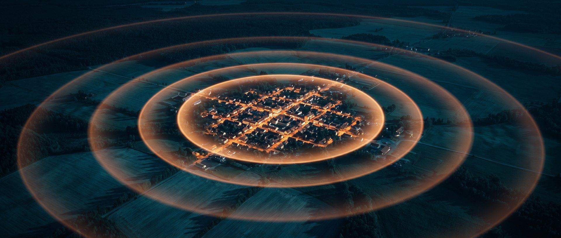 An aerial night view of a small town, its lit streets bright at the center, with concentric rings spreading outward across dark farmland and fading as they travel away from it.