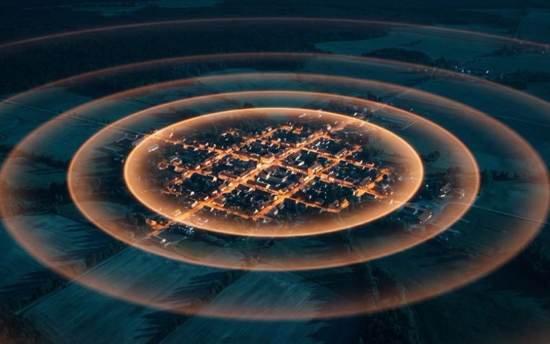 An aerial night view of a small town, its lit streets bright at the center, with concentric rings spreading outward across dark farmland and fading as they travel away from it.