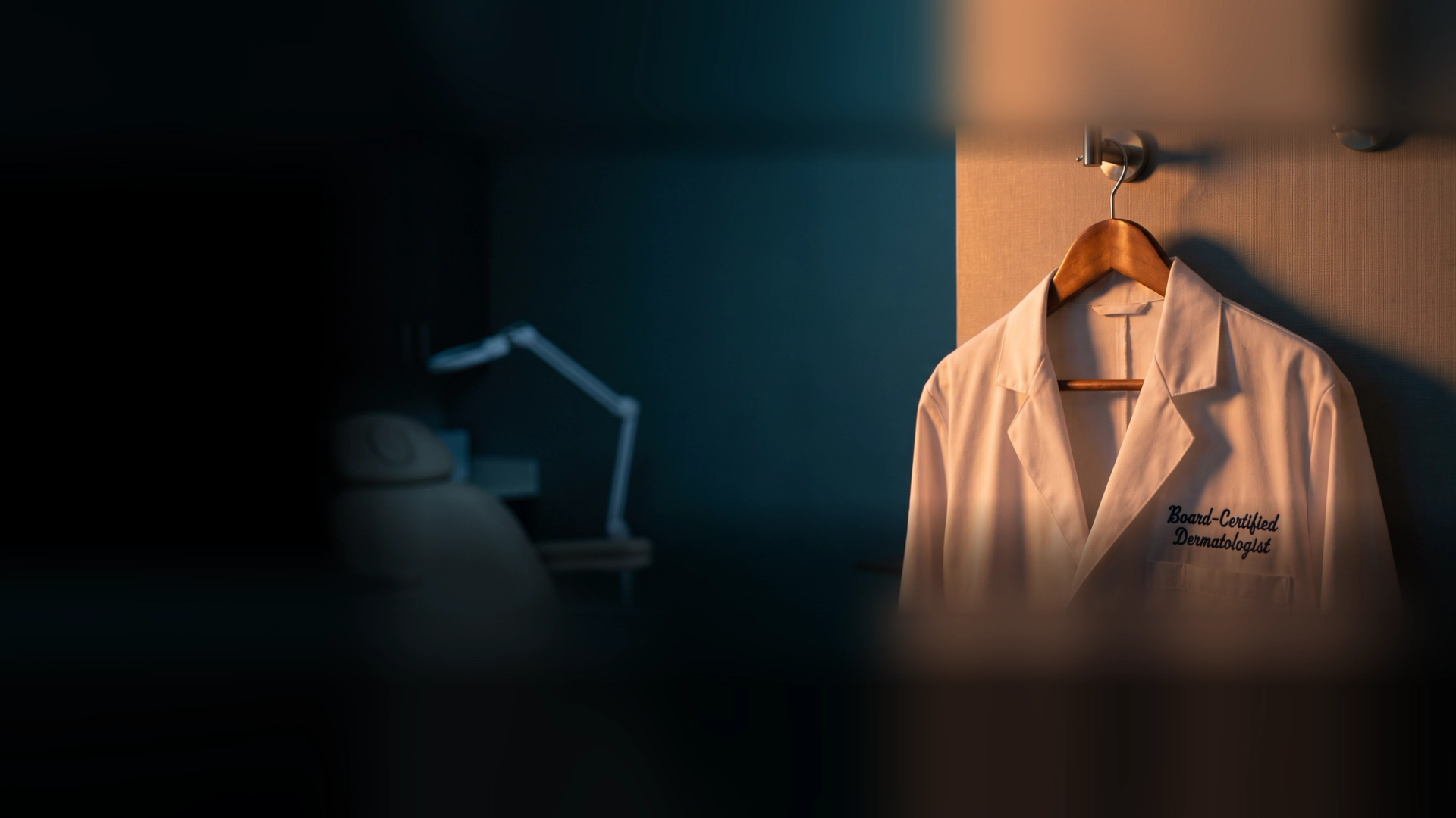 A white physician's coat on a wooden hanger, embroidered Board-Certified Dermatologist, in a dermatology treatment room