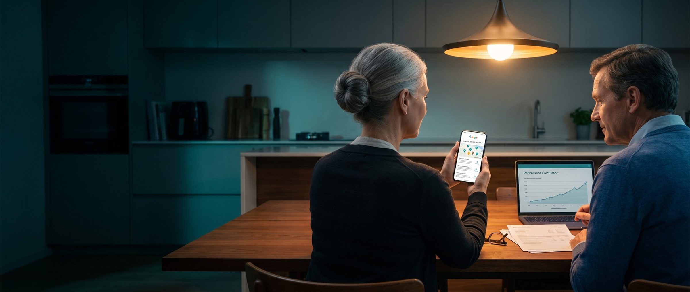 A couple in their sixties at a kitchen table at night under a pendant lamp: she holds a phone showing Google results for financial advisor near me with one amber map pin, while he sits by a laptop open to a retirement calculator chart