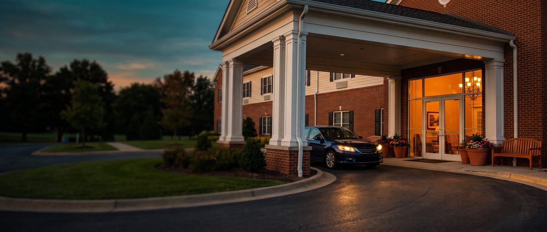 A family sedan pulled under the white-columned covered entrance of a brick senior living community at golden hour, headlights on, the lobby glowing warm through glass doors beside flower planters and a wooden bench