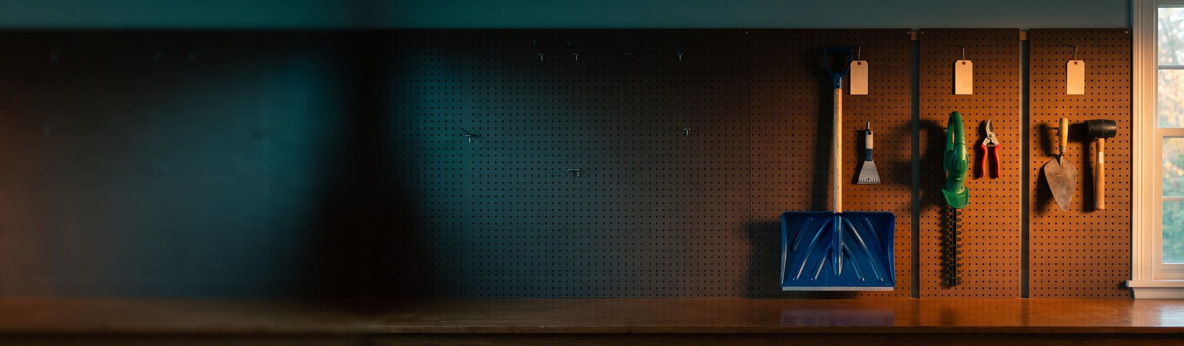 A garage pegboard wall in morning light with three separate tool groups under blank tags: a blue snow shovel and ice scraper, a green hedge trimmer and red pruners, and a mason's trowel and rubber mallet