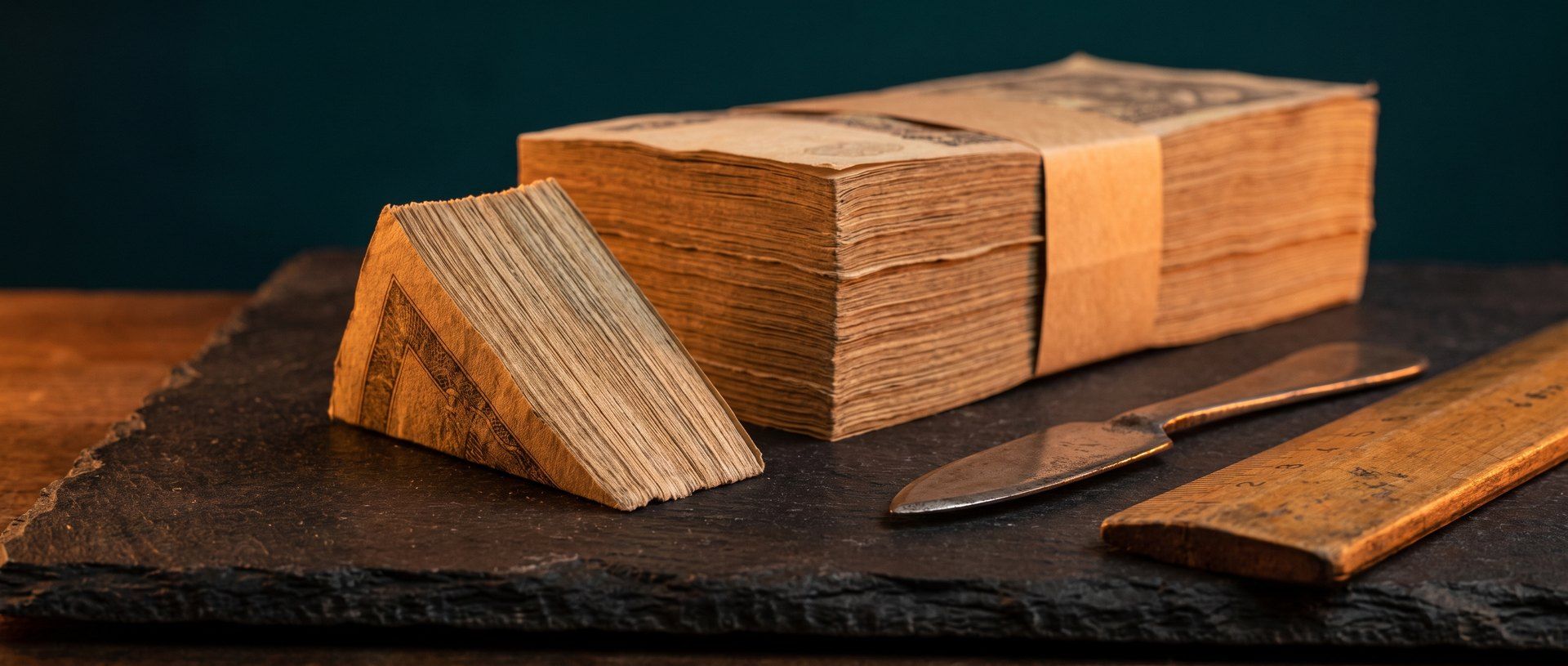 A banded stack of banknotes with a wedge cut cleanly out of it, beside a blade and a rule