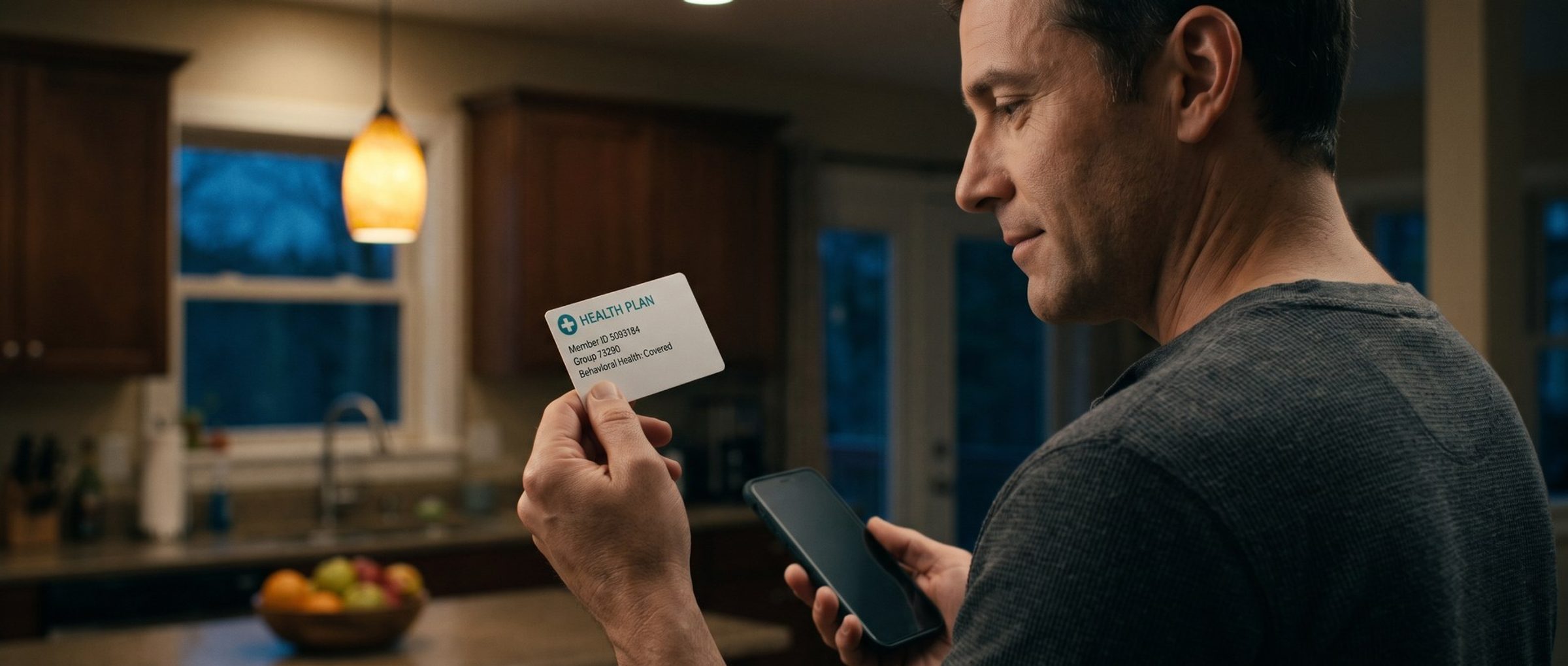 In a warm kitchen on a weekday evening, a man holds up his health insurance card, which reads Health Plan, Member ID, Group and Behavioral Health: Covered, with his phone in his other hand