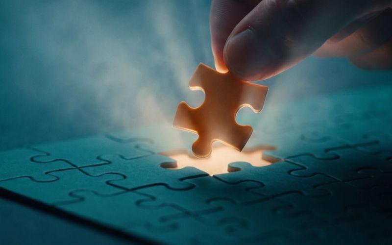 Fingertips lowering a single warm-amber puzzle piece into the last empty gap of a deep teal jigsaw, the moment a ready buyer confirms the thing actually fits.
