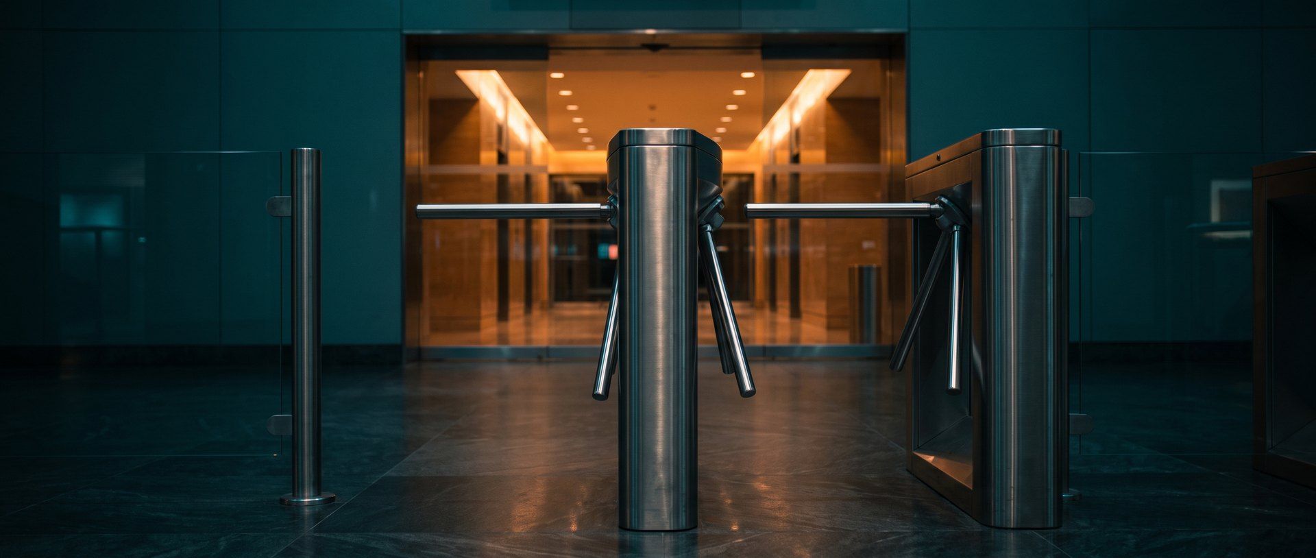 A locked steel turnstile in a dark concourse, with a warm amber lit hall beyond it that cannot be reached