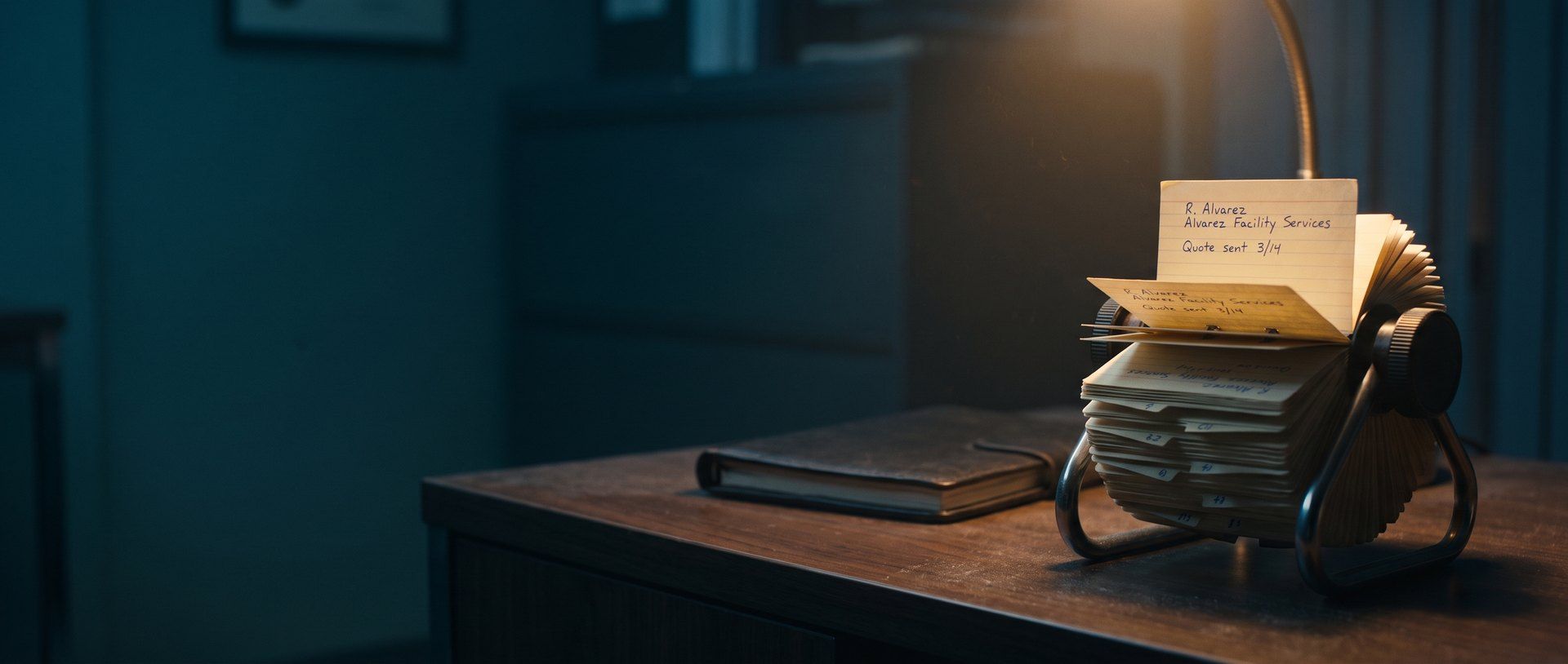 A dusty desk Rolodex in a dark office with one old contact card pulled up under a lamp, handwritten R. Alvarez, Alvarez Facility Services, Quote sent 3/14