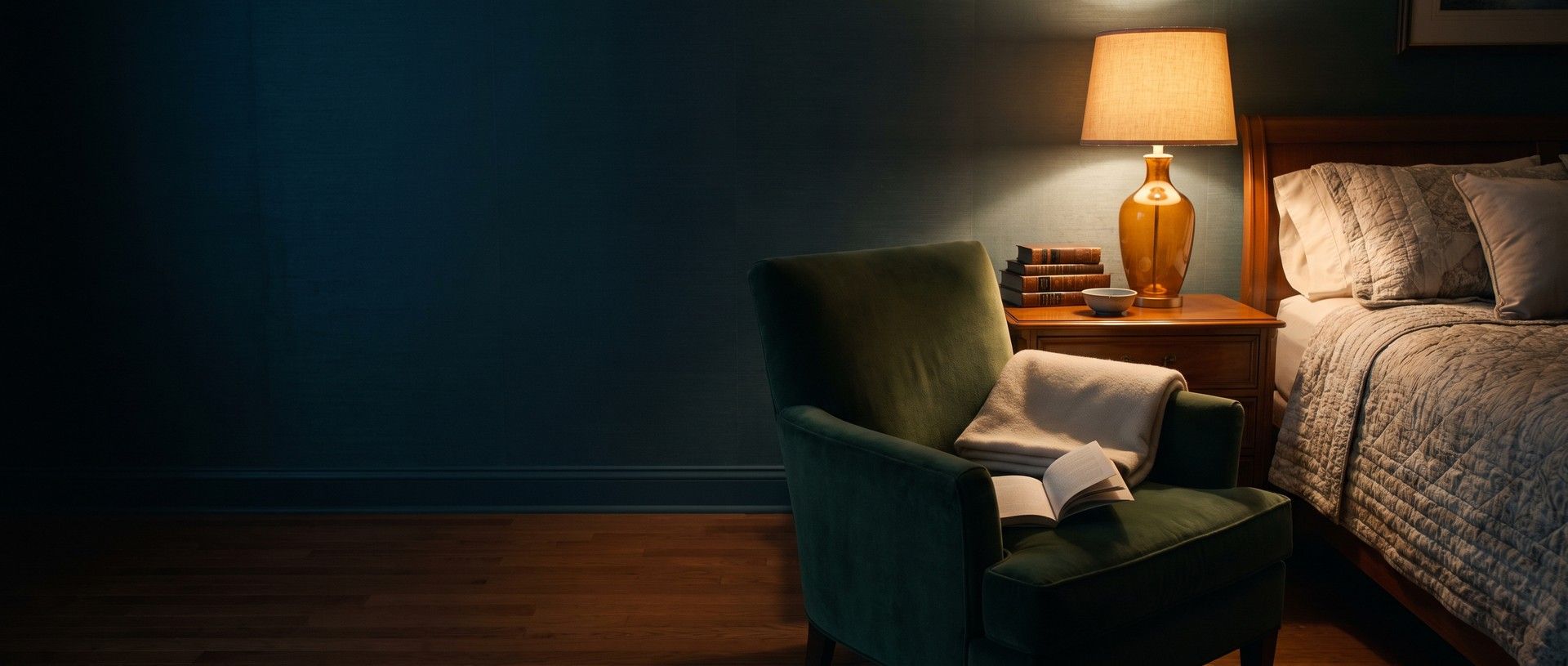 A green armchair pulled up beside a made bed at night, a folded blanket and an open book on the chair, lit by a single amber bedside lamp