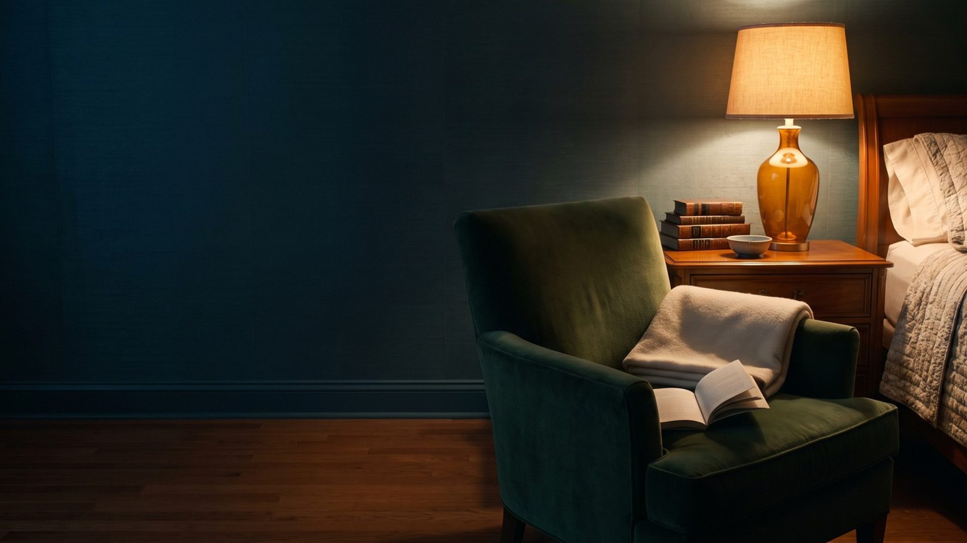 A green armchair pulled up beside a made bed at night, a folded blanket and an open book on the chair, lit by a single amber bedside lamp