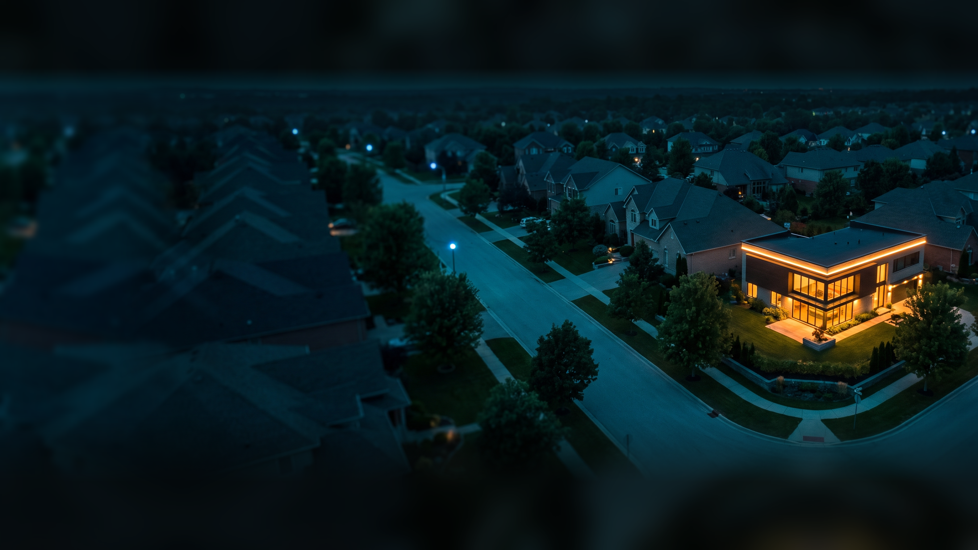 An aerial view of a quiet neighborhood at dusk, streets and rooftops in shadow, one modern building on a corner glowing warm amber