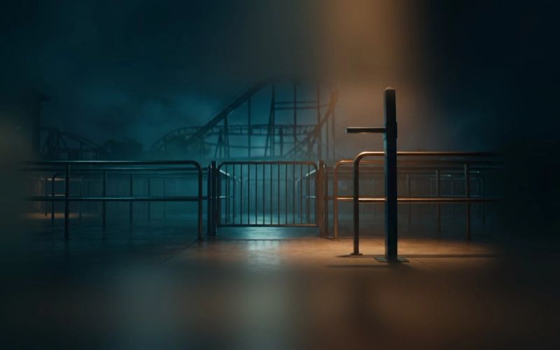 Wide view of an empty amusement park ride entrance at night, a height-check post with its measuring arm at child height lit in warm amber beside the line rails and gate, a roller coaster fading into teal fog behind