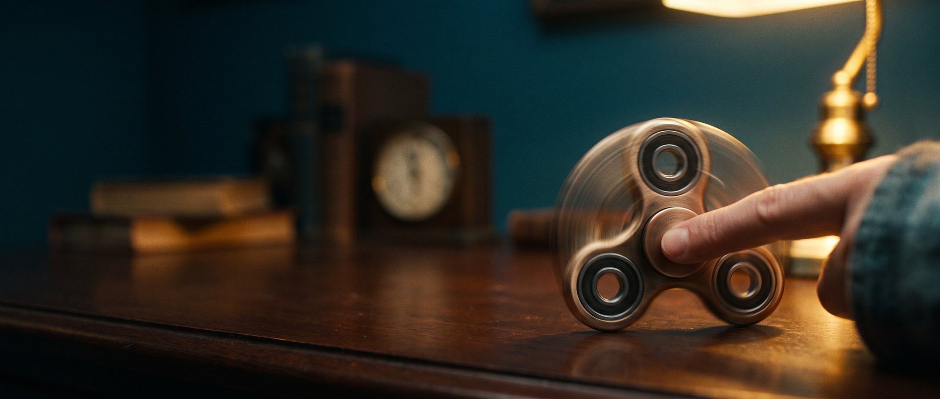 A metal fidget spinner spinning under a fingertip on a dark desk, lit warm amber against deep teal shadow
