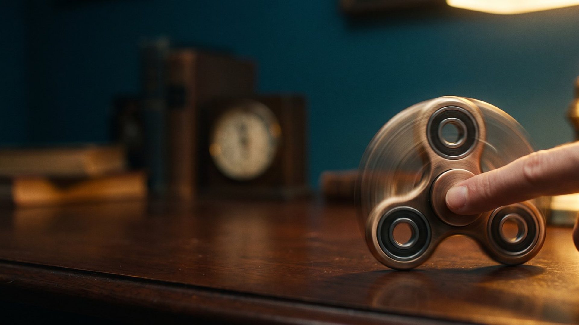 A metal fidget spinner spinning under a fingertip on a dark desk, lit warm amber against deep teal shadow