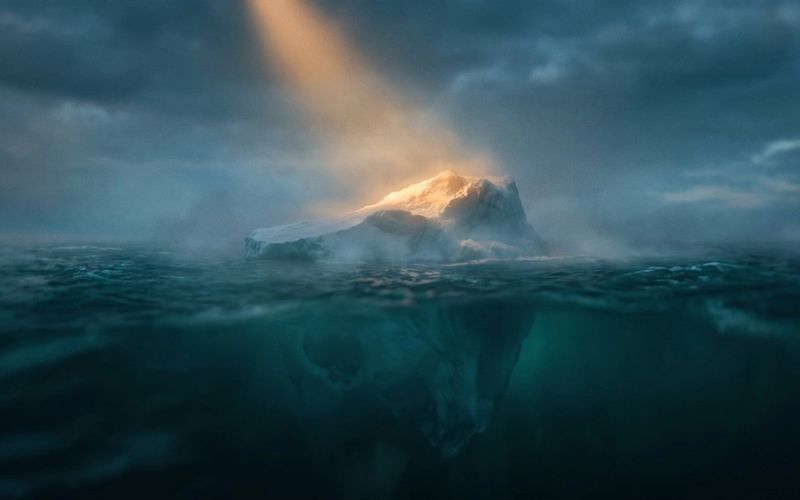 A lone iceberg at dusk, its small amber-lit tip above dark teal water and a vast shadowed mass hidden below the surface - the visible number versus the hidden reason your analytics can't show you.