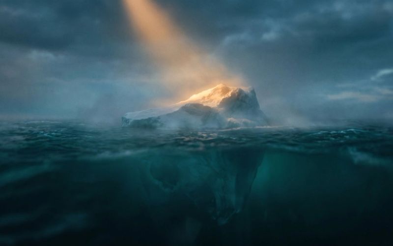 A lone iceberg at dusk, its small amber-lit tip above dark teal water and a vast shadowed mass hidden below the surface - the visible number versus the hidden reason your analytics can't show you.